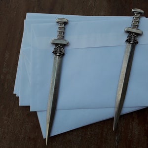 Puede incluir: Dos abridores de cartas plateados con dise&ntilde;o de espada descansan sobre una pila de sobres blancos. Los abridores tienen mangos detallados y una hoja puntiaguda. El fondo es una superficie de madera.