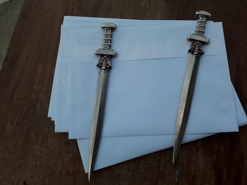 Puede incluir: Dos abridores de cartas plateados con dise&ntilde;o de espada descansan sobre una pila de sobres blancos. Los abridores tienen mangos detallados y una hoja puntiaguda. El fondo es una superficie de madera.