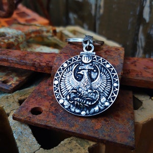 May include: Silver key chain with a round charm featuring a detailed design of a mythical bird with spread wings. The charm has a textured border and a Roman numeral III in the centre.