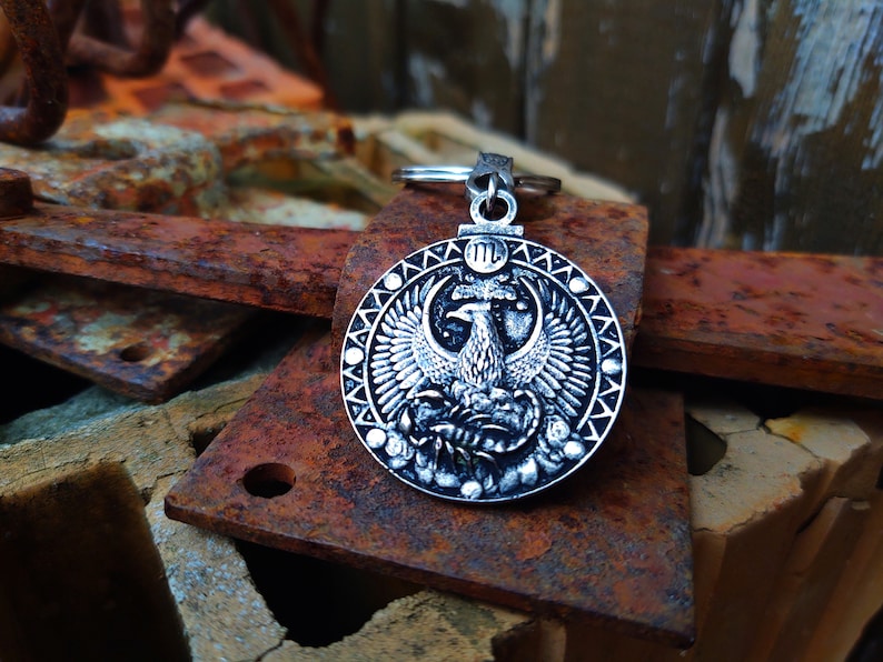 May include: Silver key chain with a round charm featuring a detailed design of a mythical bird with spread wings. The charm has a textured border and a Roman numeral III in the centre.