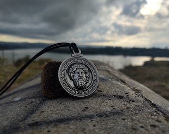 May include: A silver pendant necklace with a detailed image of a man's face surrounded by a Greek key design. The necklace is on a gray rock with a blurry background of a body of water and a cloudy sky.