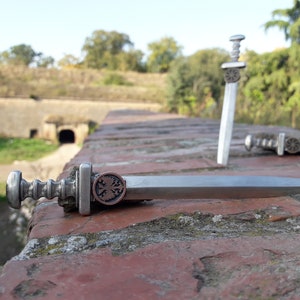May include: Two miniature silver swords with intricate details, one lying on a brick surface and the other standing upright. The sword lying down has a copper-colored emblem with a cross and two birds.