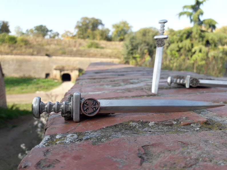 May include: Two miniature silver swords with intricate details, one lying on a brick surface and the other standing upright. The sword lying down has a copper-colored emblem with a cross and two birds.