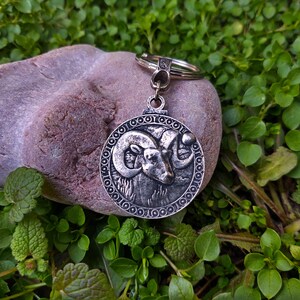 May include: Silver key chain with a round charm featuring a detailed ram head design. The charm is surrounded by a decorative border of circles. The keychain is resting on a grey rock.