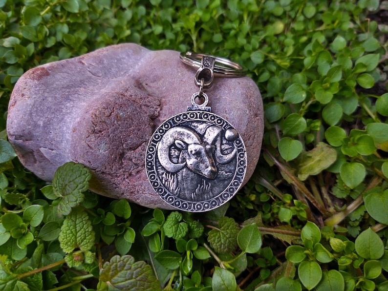 May include: Silver key chain with a round charm featuring a detailed ram head design. The charm is surrounded by a decorative border of circles. The keychain is resting on a grey rock.