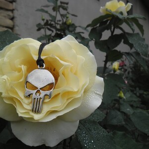 Pode incluir: Um colar com um pingente de caveira de prata com um cord&atilde;o preto pendurado em uma rosa branca.