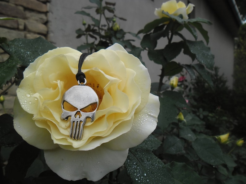 Pode incluir: Um colar com um pingente de caveira de prata com um cord&atilde;o preto pendurado em uma rosa branca.