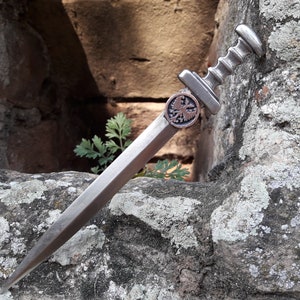 May include: A silver-colored dagger with a decorative eagle emblem on the handle. The dagger is resting on a rough, gray stone surface.
