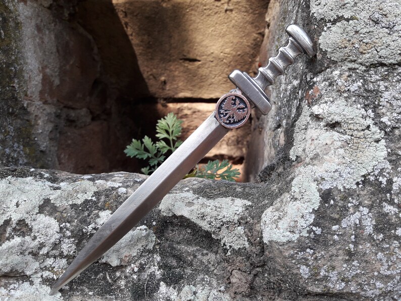 May include: A silver-colored dagger with a decorative eagle emblem on the handle. The dagger is resting on a rough, gray stone surface.