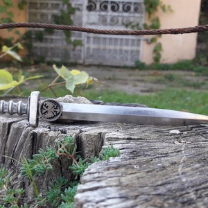 May include: A silver-toned, miniature sword with a double-headed eagle design on the handle. The sword is lying on a weathered wooden surface.