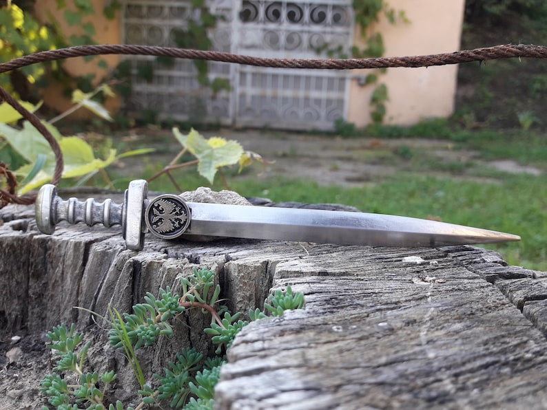 May include: A silver-toned, miniature sword with a double-headed eagle design on the handle. The sword is lying on a weathered wooden surface.