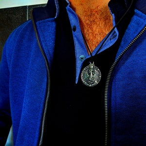 May include: A man wearing a blue jacket over a black shirt with a silver pendant necklace. The pendant features a detailed design of a bird with wings spread.