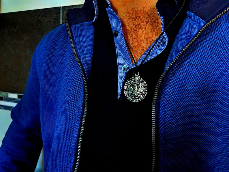 May include: A man wearing a blue jacket over a black shirt with a silver pendant necklace. The pendant features a detailed design of a bird with wings spread.