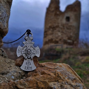 Könnte beinhalten: Ein silberfarbener Metallkreuz-Anhänger mit einem zentralen Relief eines Gesichts, kyrillischem Text und einem Kronenmotiv an der Oberseite. Der Anhänger hängt an einer dünnen schwarzen Kordel und liegt auf einer strukturierten, verwitterten Steinoberfläche.
