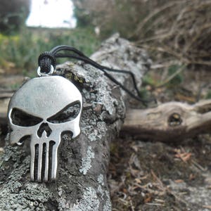 Pode incluir: Um colar com um pingente de caveira de prata com um cord&atilde;o preto. O pingente &eacute; detalhado com um rosto de caveira e uma superf&iacute;cie texturizada. O colar est&aacute; sobre um peda&ccedil;o de casca.