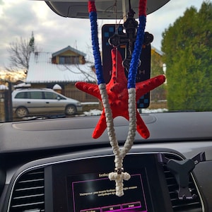 May include: A red and white crocheted starfish hanging from the rearview mirror of a car. The starfish is attached to a silver cross pendant. The car's dashboard is visible in the background, with a digital display showing the temperature and a message in Cyrillic.