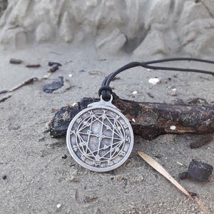 May include: A silver pendant necklace with a circular design featuring a geometric pattern and symbols. The pendant is on a black cord and is lying on a sandy surface.