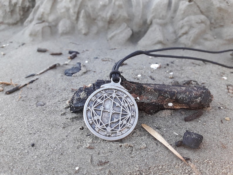 May include: A silver pendant necklace with a circular design featuring a geometric pattern and symbols. The pendant is on a black cord and is lying on a sandy surface.