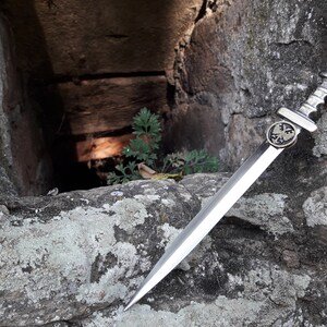 May include: A silver dagger with a black eagle emblem on the handle, resting on a rough, gray stone surface. The dagger is sharp and pointed, with a long, slender blade.