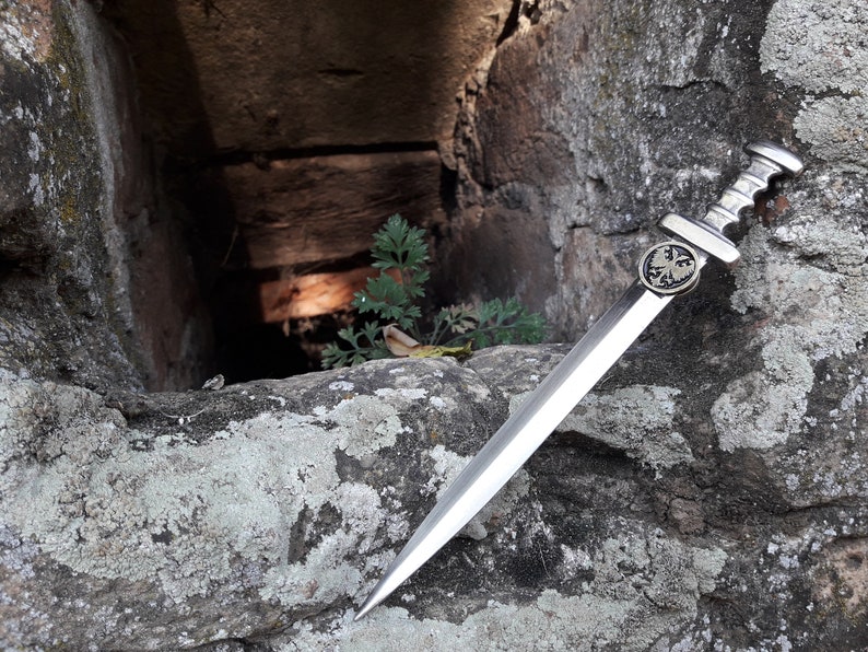 May include: A silver dagger with a black eagle emblem on the handle, resting on a rough, gray stone surface. The dagger is sharp and pointed, with a long, slender blade.