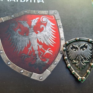 Pode incluir: Dois pinos de metal prateados em forma de escudo com um design de águia bicéfala. A águia é branca com detalhes pretos e está sobre um fundo vermelho no pino maior. O pino menor é prateado com uma águia preta.