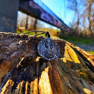 May include: A silver pendant featuring an intricate design of a mythical creature with wings, a crown, and the Virgo zodiac symbol. The pendant is on a black cord and resting on a piece of weathered wood.
