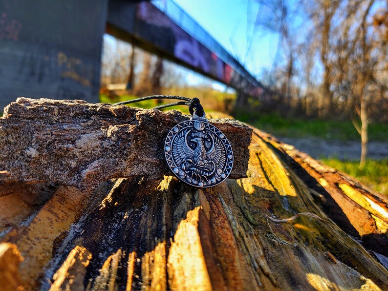 May include: A silver pendant featuring an intricate design of a mythical creature with wings, a crown, and the Virgo zodiac symbol. The pendant is on a black cord and resting on a piece of weathered wood.