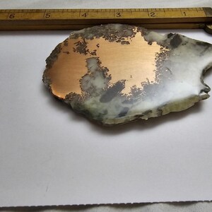 May include: A unique, irregularly shaped stone with a copper-colored metallic inlay. The stone features a mix of white, gray, and green hues, with a rough, natural edge. A wooden ruler is partially visible in the background.