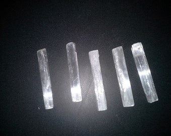 May include: Six selenite wands, also known as selenite sticks, are arranged in a row on a black surface. The wands are all white and have a smooth, polished finish. They are all approximately the same size and shape.