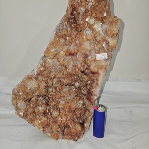 May include: A large, rough-hewn crystal formation with a mix of orange, brown, and white hues. The crystal has a textured surface and a natural, organic shape. A blue lighter is placed next to the crystal for scale.