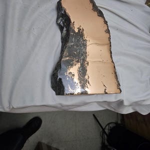 May include: A unique, irregularly shaped piece of copper-colored metal with a dark, textured border. The metal has a polished, reflective surface, and the edges are rough and uneven. The piece appears to be a decorative item or a component of a larger artwork.