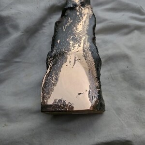 May include: A vertical, elongated sculpture featuring a rough, dark, textured exterior surface that contrasts with a smooth, polished, reflective metallic copper-colored face. The object has an irregular, organic shape with a flat base for stability.