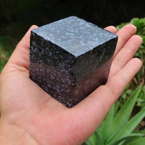 May include: A dark blue and white speckled cube of polished obsidian, approximately 2 inches in size, held in a person's hand.