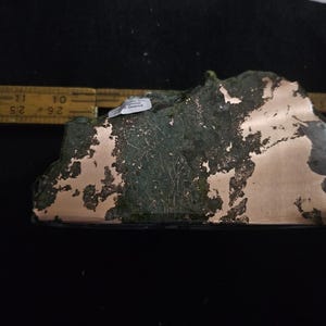 May include: A unique, irregularly shaped object with a copper-colored surface and a green, textured overlay. The object is approximately 3 inches long and appears to be a decorative piece or sculpture. The object is on a black surface.