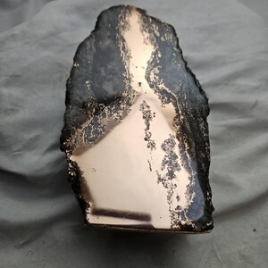 May include: A vertical, irregularly shaped stone slab featuring a polished, reflective rose gold metallic surface on the front face. The edges of the stone are dark, textured, and rough, contrasting with the smooth, mirror-like metallic finish.