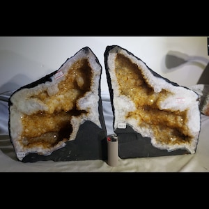 Op de afbeelding: Twee grote, natuurlijke geodehelften met een witte buitenkant en een goudbruin interieur. De geoden worden getoond op een zwart oppervlak.