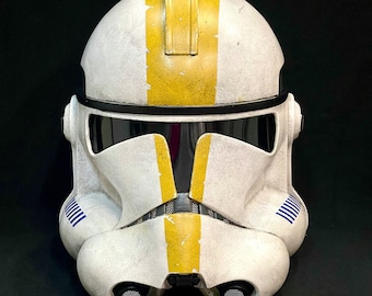 May include: A white and yellow Star Wars clone trooper helmet with blue stripes. The helmet has a weathered look and a yellow stripe down the center.