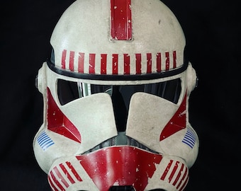 May include: A white and red clone trooper helmet with blue accents. The helmet has a weathered look and is made of plastic.