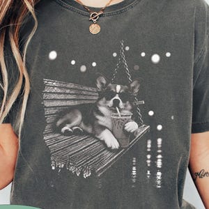 May include: A charcoal gray short-sleeve t-shirt featuring a graphic print of a corgi wearing sunglasses, lying on a wooden swing while holding a drink with a straw. The design includes white circular light accents and a green oval label with the text PEPPER.