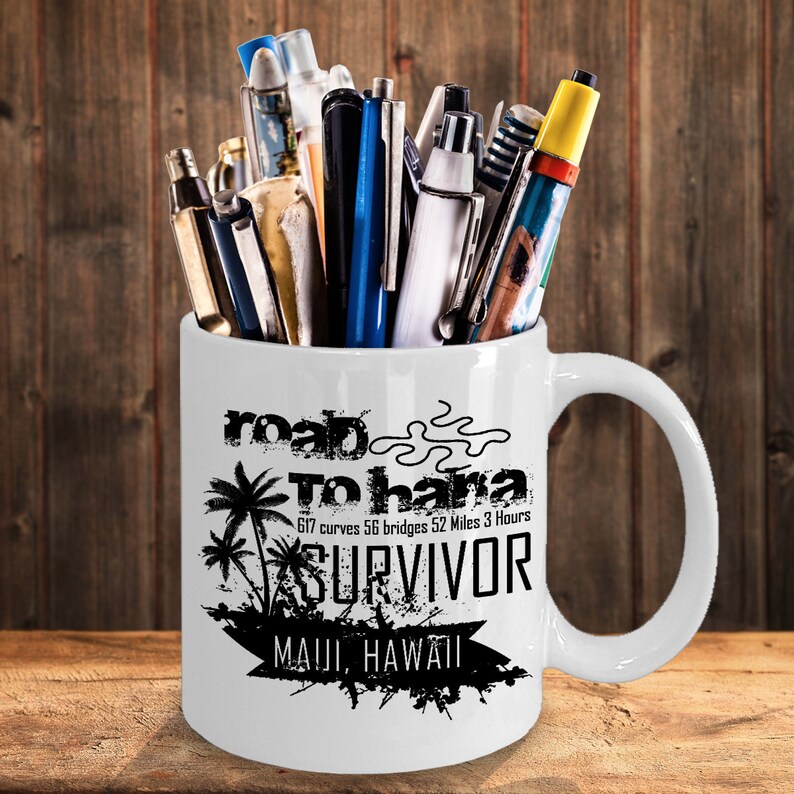 K&ouml;nnte beinhalten: Wei&szlig;e Keramiktasse gef&uuml;llt mit Stiften und Markern. Die Tasse hat einen schwarzen Text mit der Aufschrift "road To hana SURVIVOR MAUI, HAWAII" mit Palmen-Grafiken. Die Tasse steht auf einer Holzoberfl&auml;che.