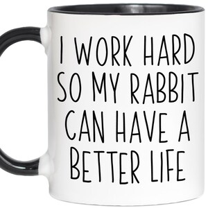 May include: White ceramic mug with a black handle and rim. The mug features the text "I WORK HARD SO MY RABBIT CAN HAVE A BETTER LIFE" in black, bold letters. A fun gift for rabbit owners.
