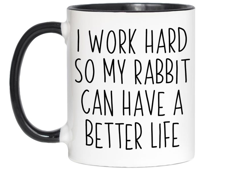 May include: White ceramic mug with a black handle and rim. The mug features the text "I WORK HARD SO MY RABBIT CAN HAVE A BETTER LIFE" in black, bold letters. A fun gift for rabbit owners.