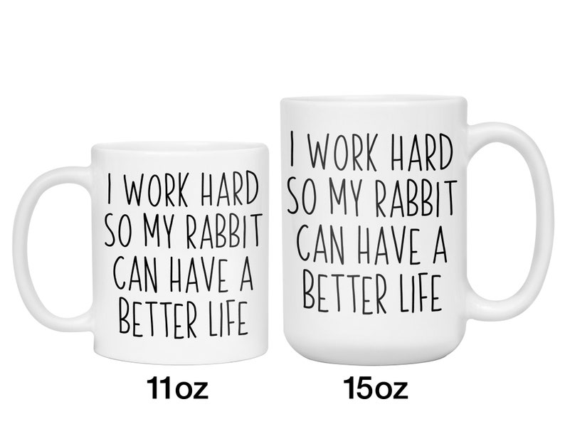 May include: Two white ceramic mugs with black text that reads "I WORK HARD SO MY RABBIT CAN HAVE A BETTER LIFE." The mugs are labeled 11oz and 15oz. The mugs have a handle on the right side.