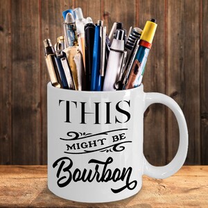 Puede incluir: Taza de cer&aacute;mica blanca con el texto "THIS MIGHT BE Bourbon" en escritura negra. La taza est&aacute; llena de bol&iacute;grafos y rotuladores de varios colores y dise&ntilde;os. La taza tiene un asa grande y est&aacute; sobre una superficie de madera.