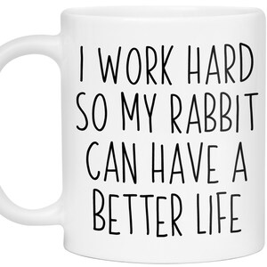 May include: White ceramic mug with the text "I WORK HARD SO MY RABBIT CAN HAVE A BETTER LIFE" in black, sans-serif font. The mug has a curved handle and is designed for hot beverages.