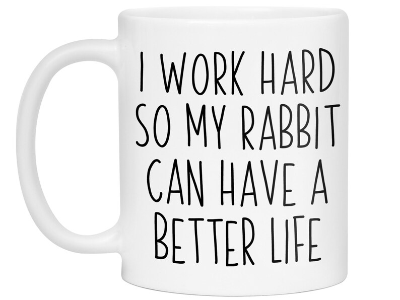 May include: White ceramic mug with the text "I WORK HARD SO MY RABBIT CAN HAVE A BETTER LIFE" in black, sans-serif font. The mug has a curved handle and is designed for hot beverages.