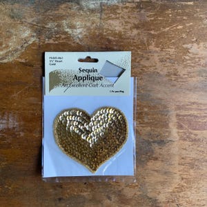 May include: A gold sequin heart applique, 3 1/2 inches in size, packaged on a white card with the text "Sequin Applique An Excellent Craft Accent 1 Pc per-Pkg."