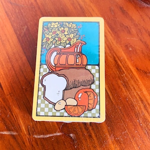 May include: A vintage-style playing card featuring a still life illustration. The image includes a brown loaf of bread, a sliced orange, a pitcher, and a bouquet of yellow flowers. The background is a checkered pattern.