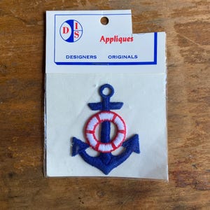 May include: A blue anchor with a red and white life preserver embroidered on it. The anchor is on a white card with the text "Appliques Designers Originals" and a logo with the letters "DIS".