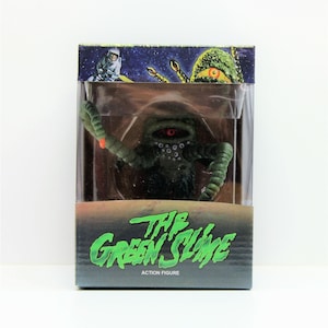 Op de afbeelding: Een groene en zwarte action figure van The Green Slime uit de film van 1968. De figuur zit in een doorzichtige plastic doos met een zwarte en groene achtergrond. De doos heeft de tekst "The Green Slime" en "Action Figure" erop.